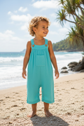A toddler with curly blonde hair stands on a sandy beach wearing a light blue linen-look overall romper. The background features the ocean, palm trees, and a bright sky.