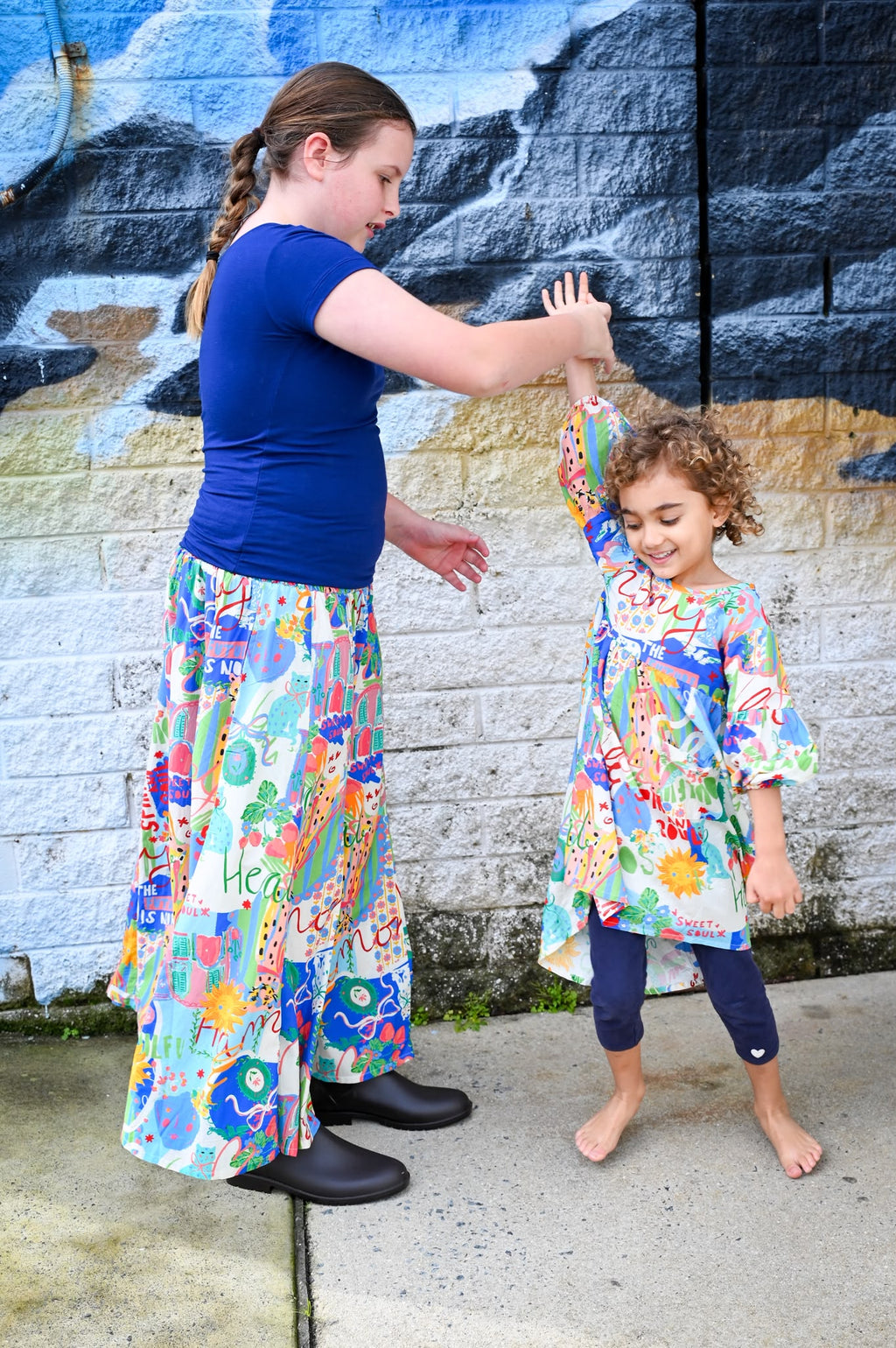 A woman and a young girl standing outdoors in front of a painted brick wall, both wearing matching colorful patterned Byron Pant and Top Sweet SoulPrint outfits.