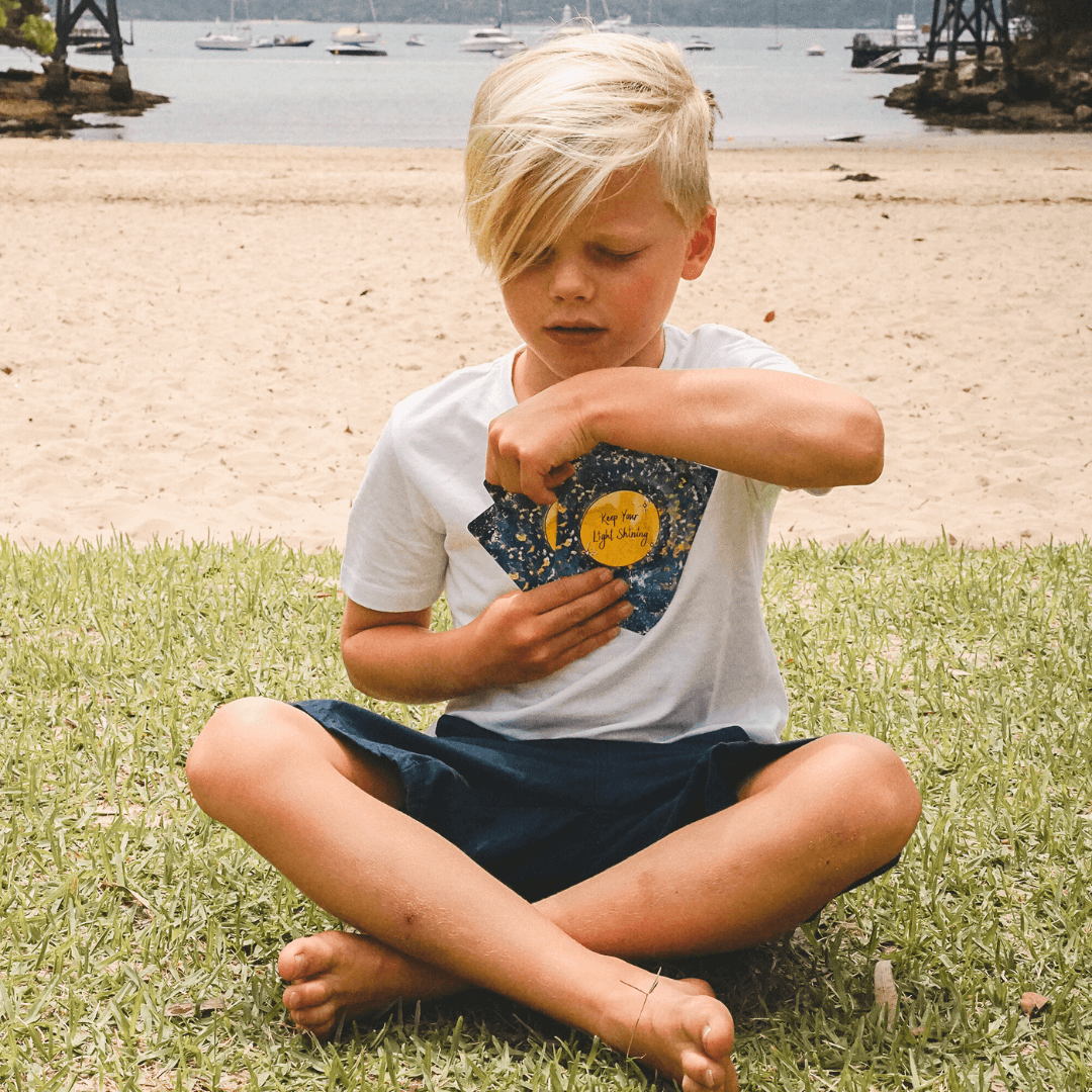 Young boy sitting on grass by a beach with boats in the background