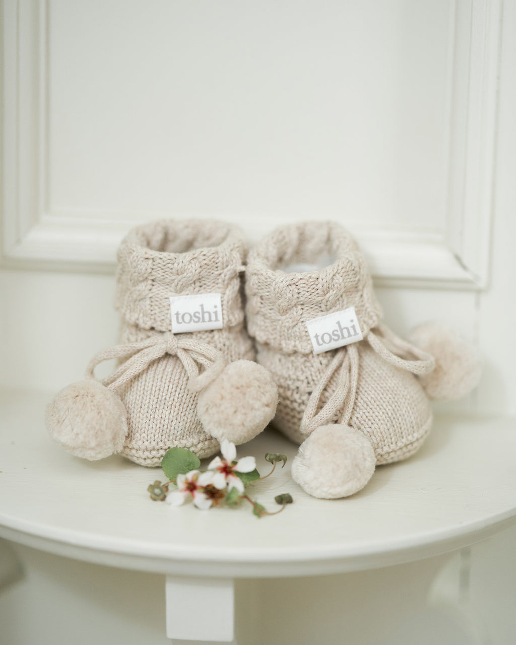 Pair of beige knitted baby booties with pom-poms on a white surface.
