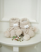 Pair of beige knitted baby booties with pom-poms on a white surface.
