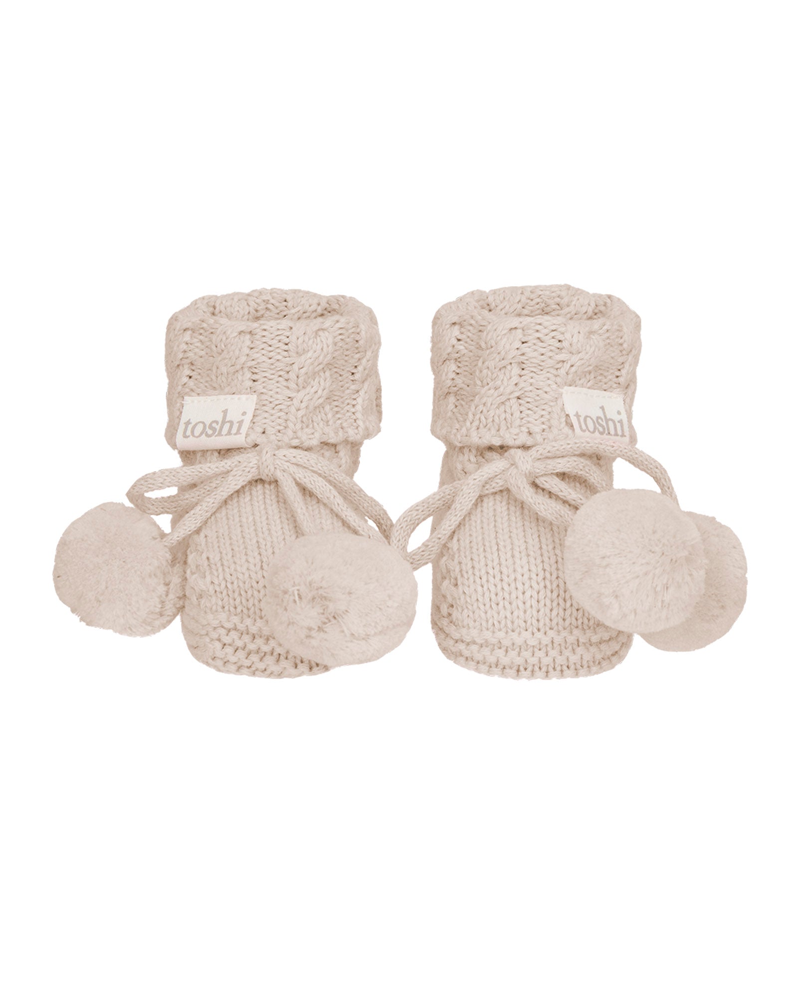 Oatmeal, a Pair of beige knitted baby booties with pom-poms on a white background.