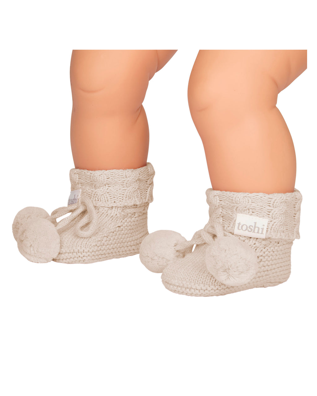 Baby's feet wearing Oatmeal, a Pair of beige knitted baby booties with pom-poms on a white background.