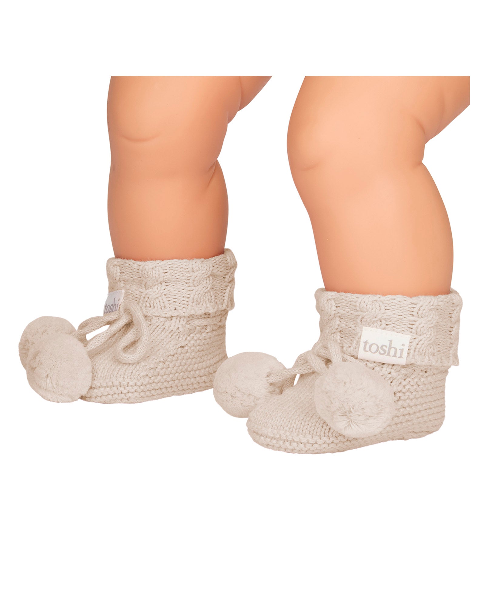 Baby's feet wearing Oatmeal, a Pair of beige knitted baby booties with pom-poms on a white background.