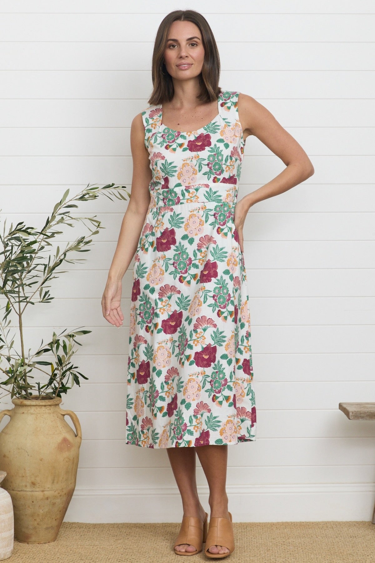 Lifestyle shot of a model wearing a knee-length summer dress featuring a botanical print of burgundy, green, and soft orange flowers against a white fabric background, accented by a wide scoop-square neckline.