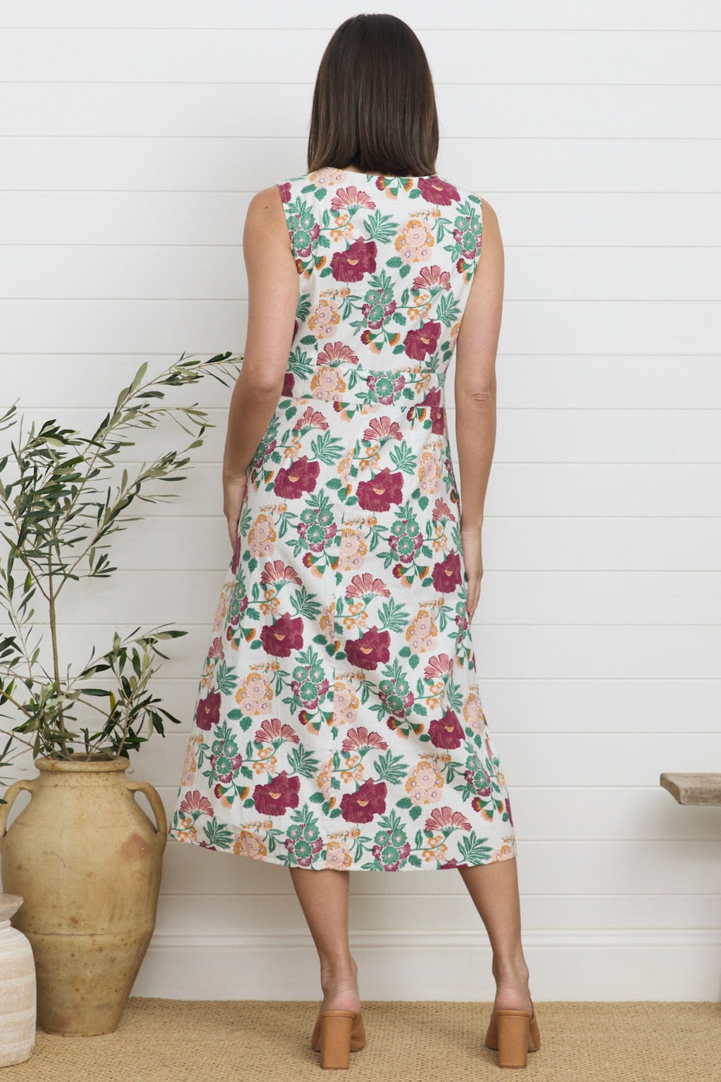 Lifestyle shot of a model wearing a knee-length summer dress featuring a botanical print of burgundy, green, and soft orange flowers against a white fabric background, accented by a wide scoop-square neckline.