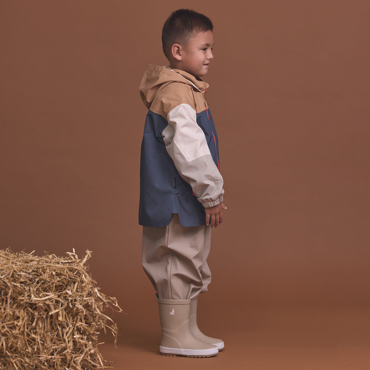 Child wearing the Crywolf Packable Jacket Ash Colourblock and pants standing next to a bale of hay on a brown background