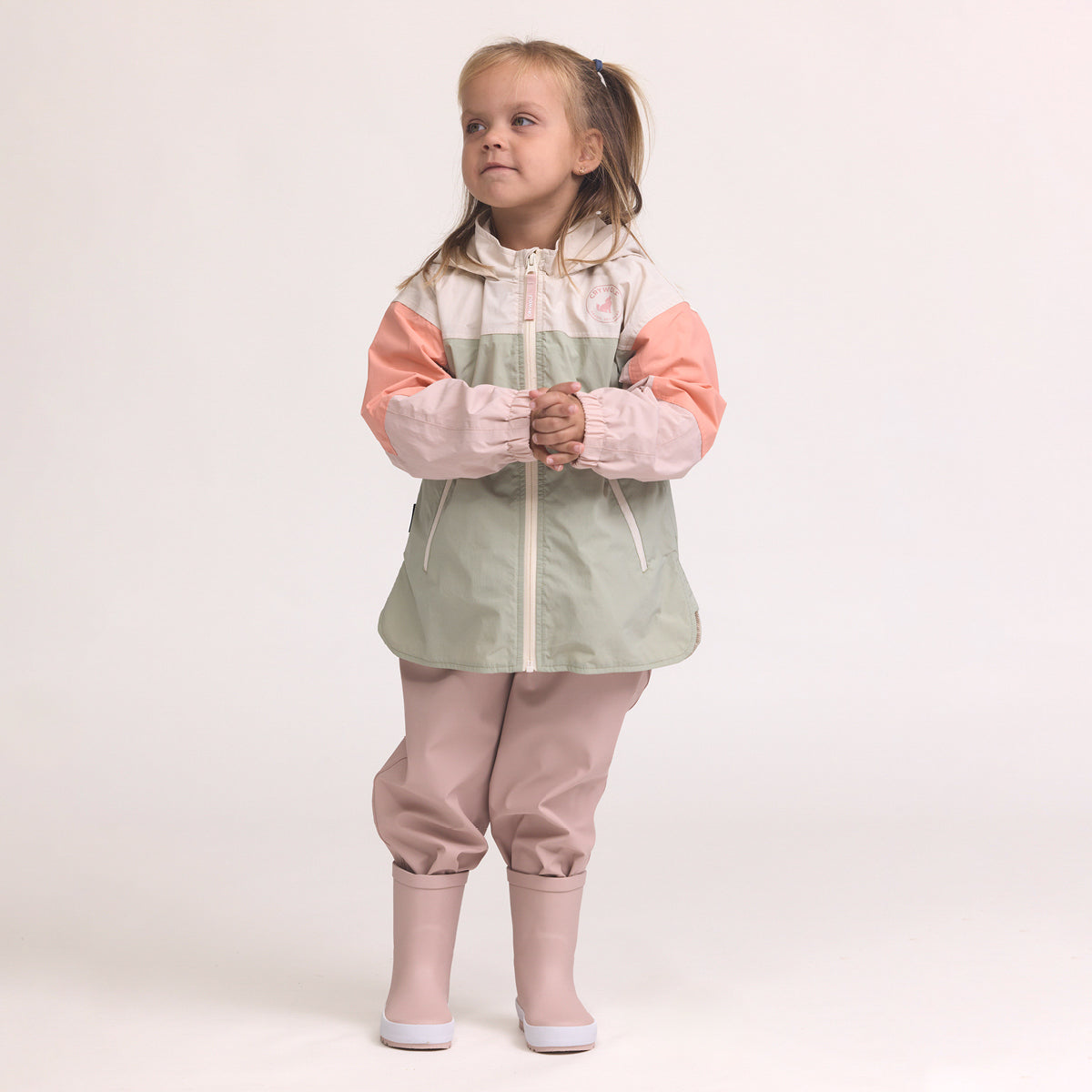 Child wearing a Color-blocked kids' waterproof jacket featuring an off-white upper body and hood, a sage green central torso, and terracotta pink sleeves with a cream zipper, pink pants and Crywolf Rain Boots Dusty Pink on a light background.