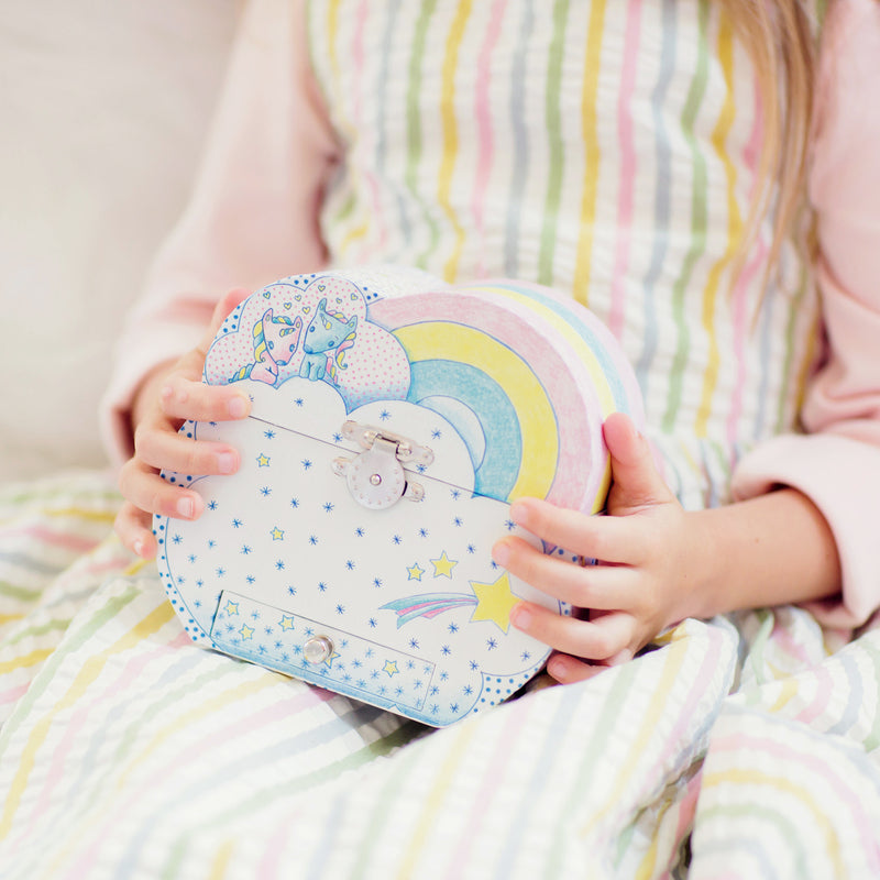 Child holding an open pastel blue children's music box decorated with yellow stars and white dots, featuring a yellow-lined interior, cloud-shaped vanity mirror, and a white unicorn figurine with a pink and blue mane.