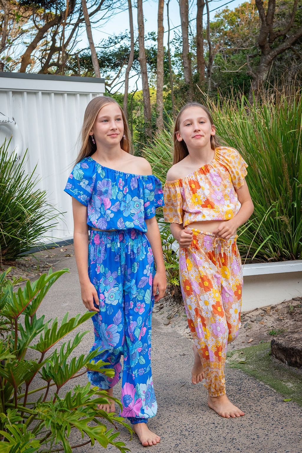 Girl in left wearing a bohemian-style outfit consisting of a blue short-sleeve crop top and matching trousers decorated with a colorful floral pattern  Lapiz Dahlia print. Girl in right in wearing two piece set of  Sun Daisy print, a vintage floral pattern in mustard yellow, rust orange, and dusty pink, featuring a smocked elastic top, adjustable shoulder ties, and wide-leg trousers.
