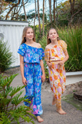Girl in left wearing a bohemian-style outfit consisting of a blue short-sleeve crop top and matching trousers decorated with a colorful floral pattern  Lapiz Dahlia print. Girl in right in wearing two piece set of  Sun Daisy print, a vintage floral pattern in mustard yellow, rust orange, and dusty pink, featuring a smocked elastic top, adjustable shoulder ties, and wide-leg trousers.