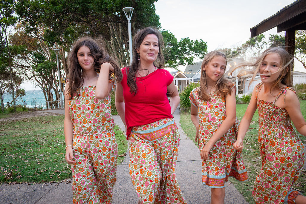 Woman with three young girls wearing Sleeveless children's jumpsuit featuring Festival Garden print, a white base with an all-over floral pattern of orange, pink, and green. 