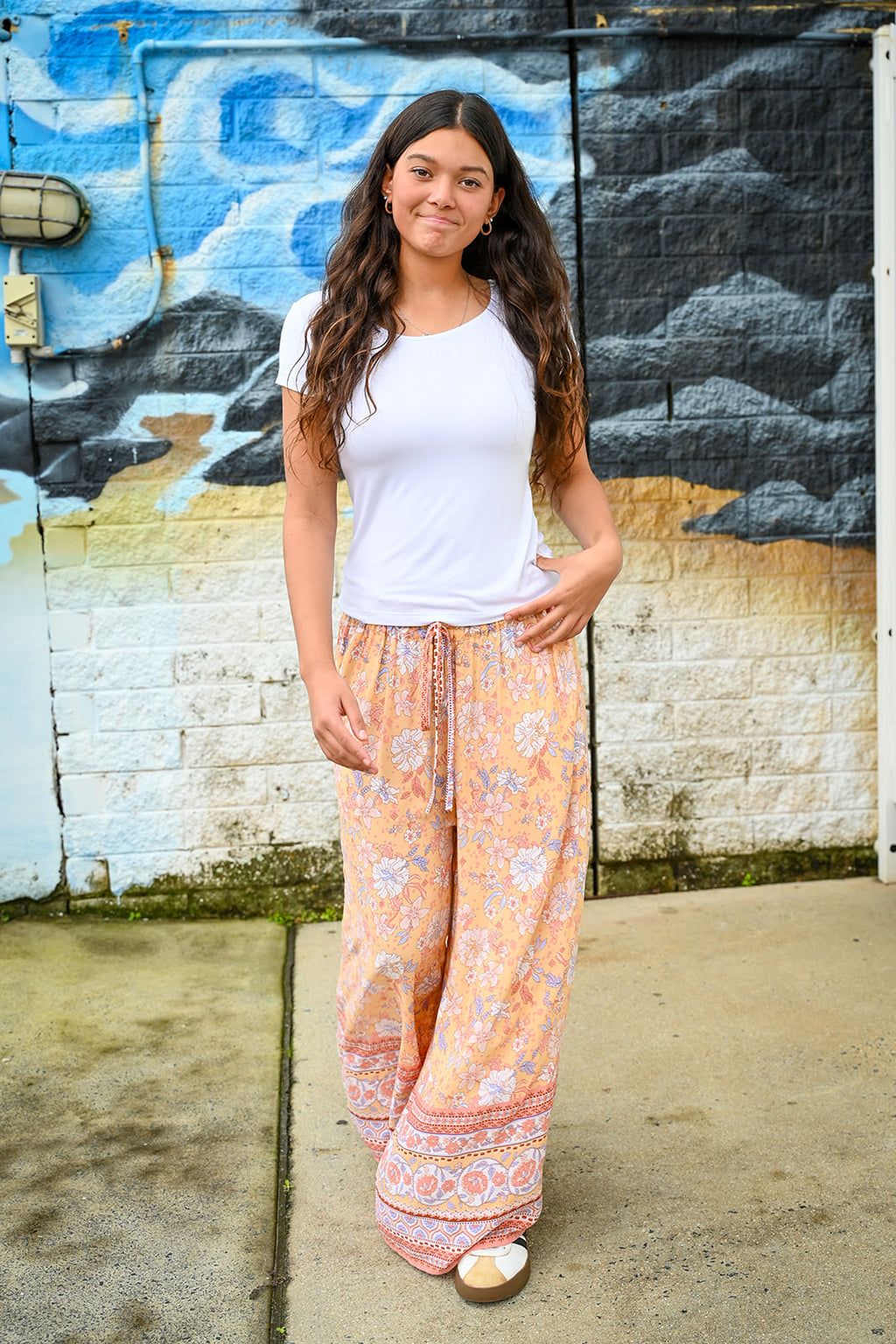 A woman stands outdoors in front of a painted brick wall, wearing loose, wide-leg peach and cream patterned pants with a white fitted T-shirt.