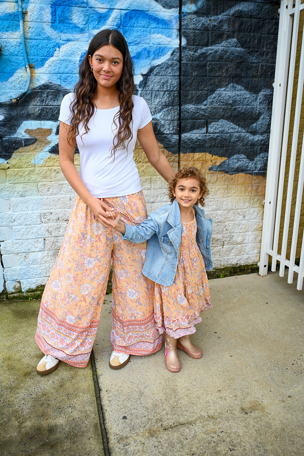 A woman and a young girl standing outdoors in front of a painted brick wall. The woman wears a white short-sleeve top and loose floral wide-leg pants, while the girl wears a light blue denim jacket over a matching floral dress.