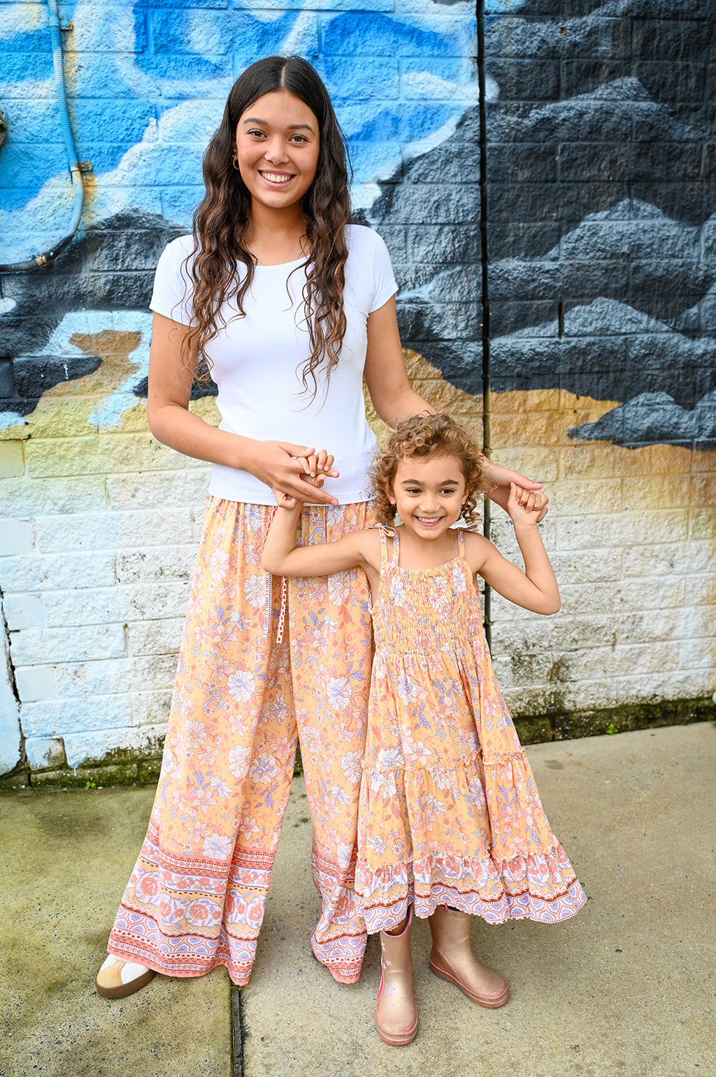 A woman and a young girl standing together in front of a painted brick wall. The woman wears a white short-sleeve top and a loose peach floral maxi skirt, while the girl wears a matching peach floral sleeveless dress.