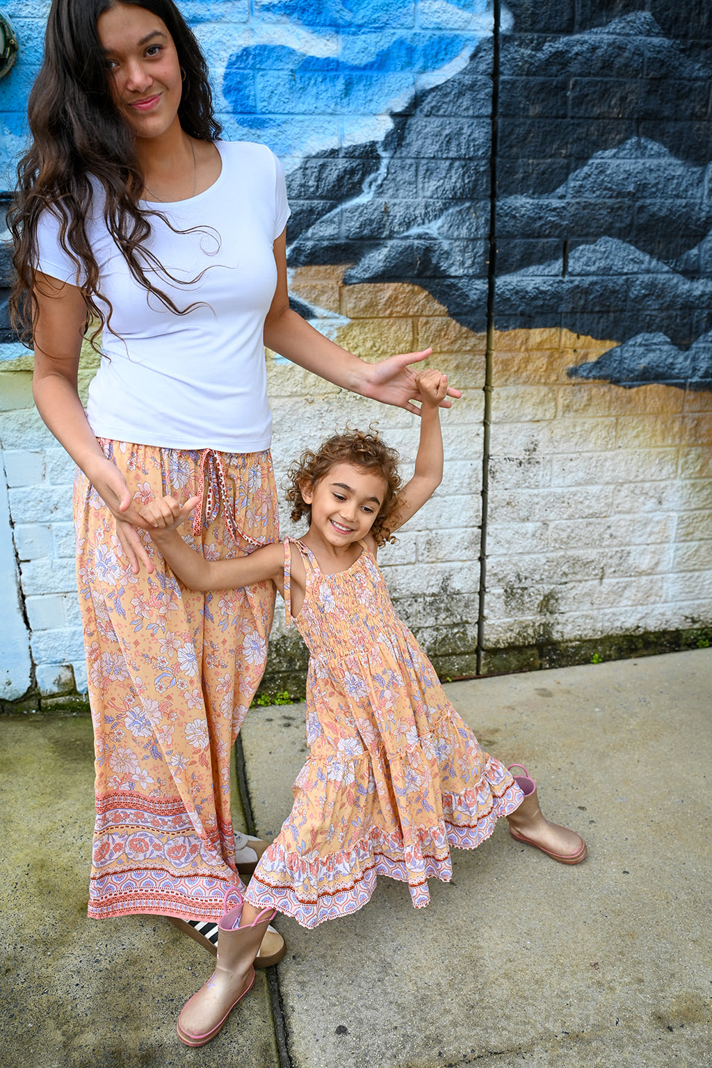 A woman and a young girl holding hands, wearing matching bohemian-style maxi dresses with a floral print in orange, pink, and blue. They are standing outdoors in front of a painted brick wall.
