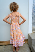 A young girl with curly brown hair is shown from behind wearing a tiered Sunrise Botanica maxi dress.