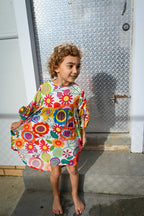 A young girl with curly brown hair stands barefoot against a weathered concrete wall, wearing a Evie Dress Flower Pop Brights.