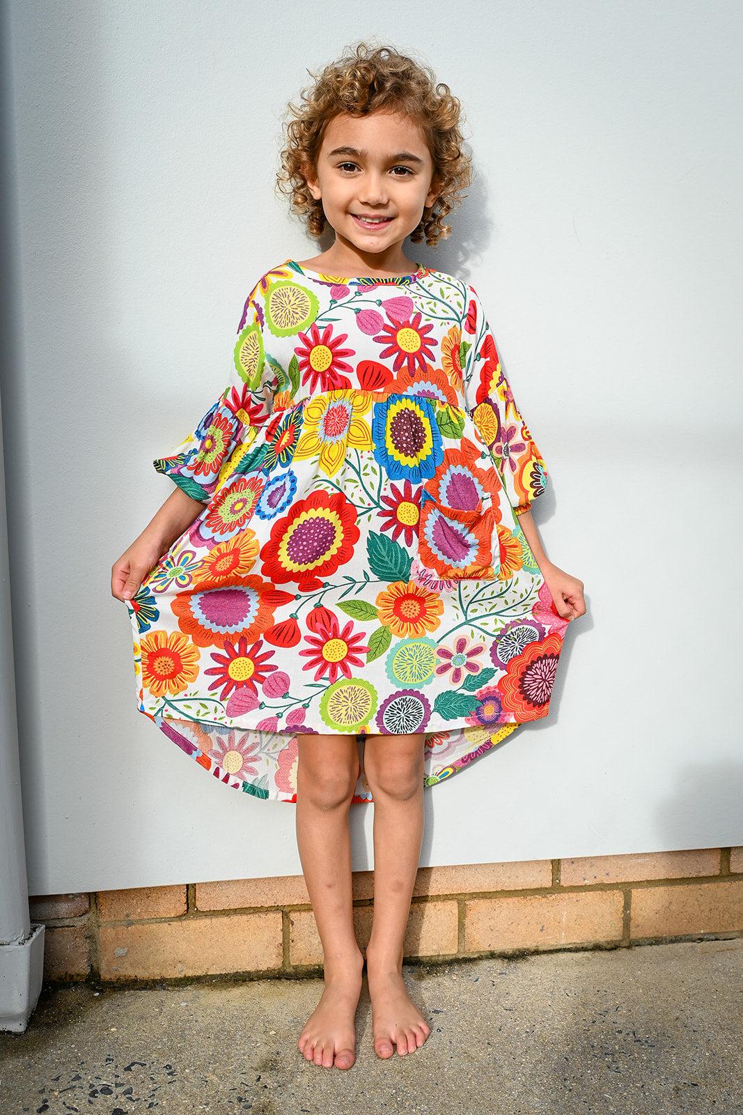 A young girl with curly brown hair stands barefoot against a weathered concrete wall, wearing a Evie Dress Flower Pop Brights.