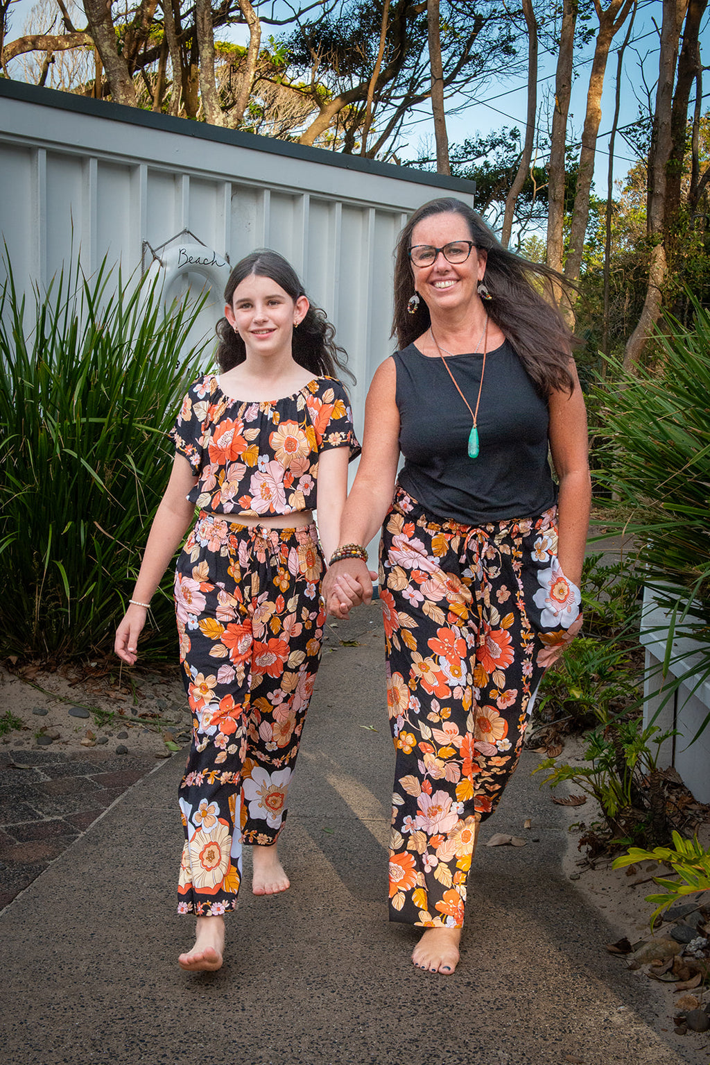 Two women wearing a black two-piece set featuring a short-sleeve top and wide-leg pants with Wild Dahlia print, a bold retro floral print in warm orange, mustard, and peach.