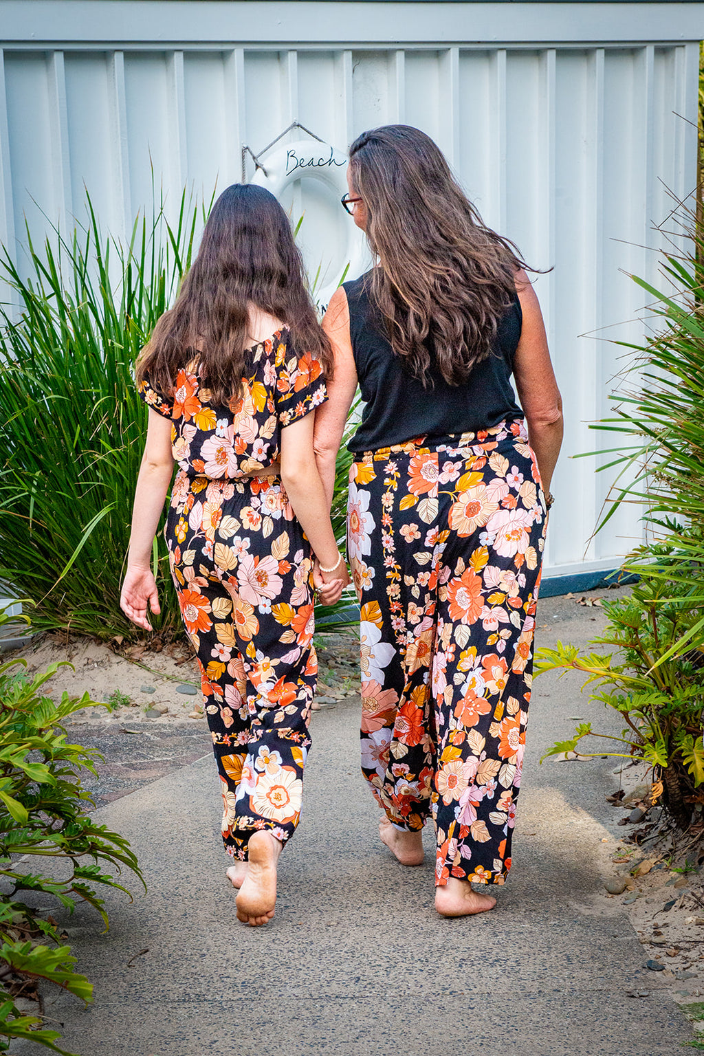 Two women walking away wearing matching black two-piece set featuring a short-sleeve top and wide-leg pants with Wild Dahlia print, a bold retro floral print in warm orange, mustard, and peach.