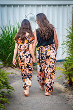 Two women walking away wearing matching black two-piece set featuring a short-sleeve top and wide-leg pants with Wild Dahlia print, a bold retro floral print in warm orange, mustard, and peach.