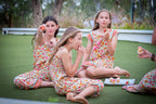 Young girls in matching Festival Garden print, a white base with an all-over floral pattern of orange, pink, and green. Sitting on grass, eating cupcakes.