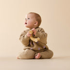 Baby in a brown knitted outfit holding a wooden toy on a beige background