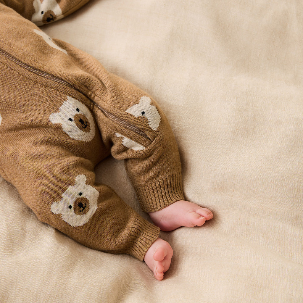 Baby wearing a Wilson & Frenchy Organic Knitted Jacquard Growsuit, a brown onesie with bear patterns while lying on a beige blanket
