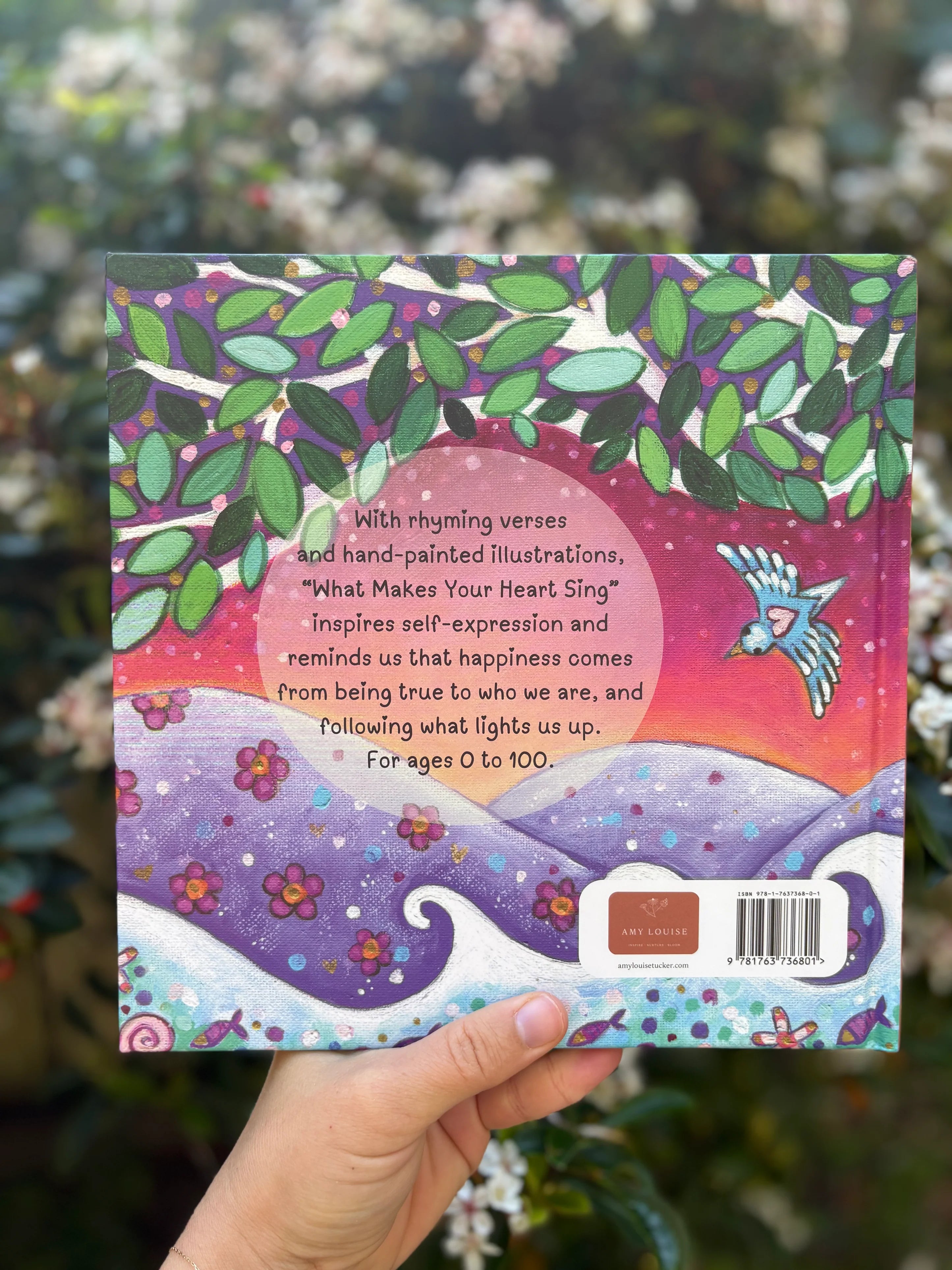 Colorful book cover with text and illustrations, held by a hand against a blurred natural background.
