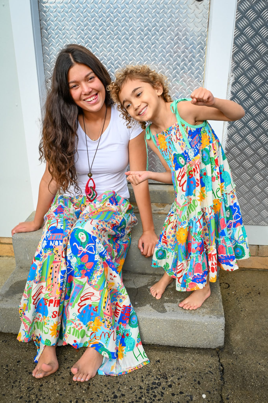 A woman and a young girl sitting together on concrete steps, wearing matching colorful abstract-patterned maxi dresses and long skirts in Byron  Sweet SoulPrint.