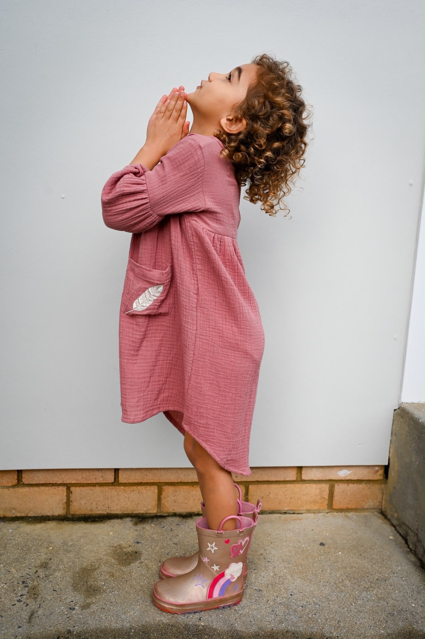 A young girl with curly brown hair is shown from the side, wearing a MilaMeli Evie Dress Dusty Rose Muslin Cotton Embroidered Feather Pocket.
